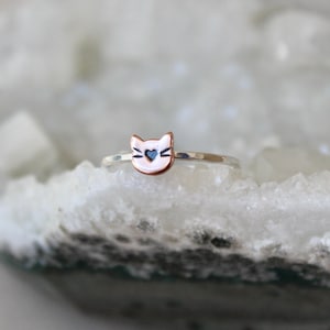 May include: A silver ring with a cat face design. The cat face is copper-colored with a blue heart in the center. The ring is set against a white, textured background.