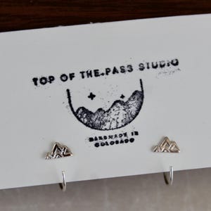 May include: A pair of silver mountain-shaped stud earrings attached to a white display card. The card features the black printed text TOP OF THE PASS STUDIO, a graphic of mountains with stars, and the text HANDMADE IN COLORADO.
