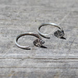 May include: Two open-ended silver hoop earrings with a geometric diamond-shaped design. The earrings are displayed on a weathered, gray wooden surface, showcasing their minimalist style and metallic shine.