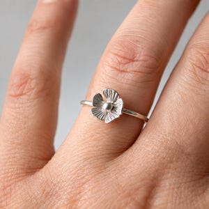 May include: A silver-toned ring featuring a small, five-petaled flower charm. The flower has a textured, radiating line pattern on each petal and a smooth, round bead at its center. The band is thin, plain, and circular in its design.
