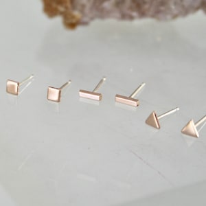 May include: A collection of six small stud earrings in various geometric shapes: square, bar, and triangle. The earrings are a rose gold color and are displayed on a white surface.