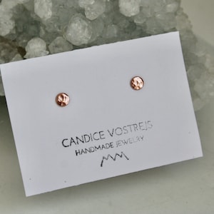 May include: A pair of small, hammered, rose gold stud earrings on a white card with the text "Candice Vostrejs Handmade Jewelry" and a mountain range logo.