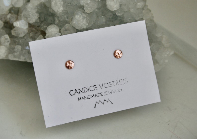 May include: A pair of small, hammered, rose gold stud earrings on a white card with the text "Candice Vostrejs Handmade Jewelry" and a mountain range logo.