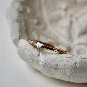 May include: A thin, polished rose gold band ring featuring a small, centered silver heart charm. The metal band has a smooth, reflective surface, and the heart is positioned prominently at the top of the ring, creating a delicate and simple jewelry piece.