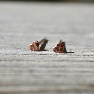 May include: A pair of small, hammered copper heart stud earrings. The earrings are on a silver post and have a textured surface. The earrings are displayed on a weathered wooden surface.