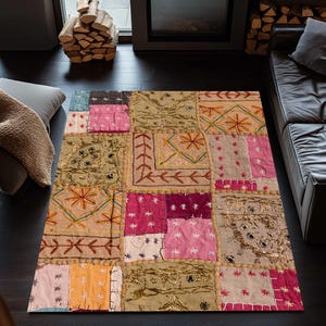 Könnte beinhalten: Ein Patchwork-Teppich mit verschiedenen Farben und Mustern. Der Teppich zeigt Quadrate aus rosa, beige und goldfarbenem Stoff mit gestickten Details. Er liegt auf einem dunklen Holzboden in einem Wohnzimmer.