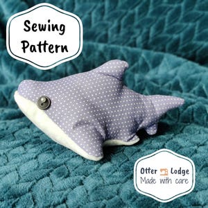 May include: A purple and white polka dot shark plush toy with a button eye. The toy is lying on a teal blue fuzzy blanket. The text "Sewing Pattern" is in a white oval on the top left of the image. The text "Otter Lodge Made with care" is in a white oval on the bottom right of the image.