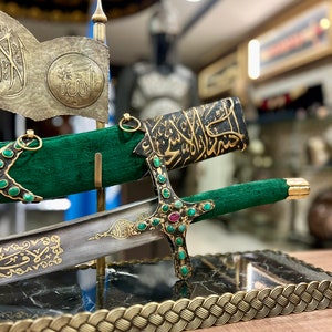 Puede incluir: Una espada decorativa con una vaina verde y dorada, un mango negro con detalles dorados y verdes, y una base de mármol negro con un ribete trenzado dorado. La espada se exhibe con una bandera de tela verde con caligrafía árabe dorada.
