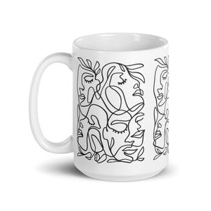 Può includere: Tazza in ceramica bianca con un disegno a linea nera di più volti e foglie. Il design è ripetuto su entrambi i lati della tazza.