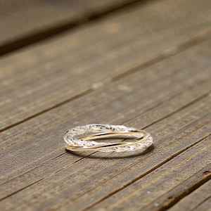 May include: A silver and gold ring with an interwoven design. The ring features a textured silver band intertwined with a smooth gold band. The ring is displayed on a rustic wooden surface.