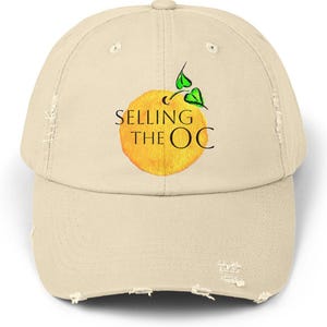 May include: A distressed beige baseball cap featuring the text "SELLING THE OC" above a watercolor-style orange with green leaves. The cap has a curved brim and a distressed, worn appearance.