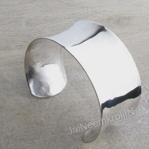 Op de afbeelding: Een brede, gepolijste zilveren manchetarmband met een gebogen ontwerp. De open armband heeft een glad, reflecterend oppervlak dat het licht opvangt. Het sieraad wordt gepresenteerd op een neutrale grijze ondergrond.