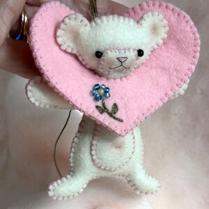 Op de afbeelding: Een handgemaakt wit vilten teddybeerornament met een roze hartvormige kraag. De beer heeft zwarte ogen, een roze neus en een kleine blauwe bloem met groene bladeren. Het ornament wordt vastgehouden door een groen touwtje.