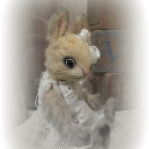 May include: A 3.25" tall, cream-colored bunny wearing a white lace dress and a white bow. The bunny is sitting on a white surface with a brick wall in the background. The text "3.25" Genevieve" is written above the bunny. The text "www.BoohBears.com" is written below the bunny.