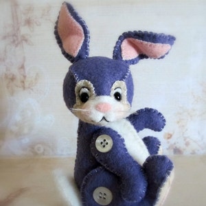 May include: A purple felt bunny with white felt accents and button eyes. The bunny is sitting with its legs crossed and has a white felt tail.