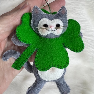 Può includere: Ornamento a forma di gatto in feltro fatto a mano con un design a trifoglio verde. Il gatto grigio ha dettagli bianchi, occhi neri e un naso rosa. L'ornamento è cucito con filo bianco e ha un cordino verde per appenderlo.