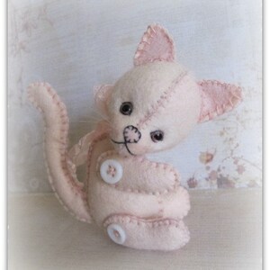 Könnte beinhalten: Ein rosa Filzkatzenspielzeug mit gestickten Details und Knopfaugen. Die Katze sitzt auf einer hellbraunen Holzoberfläche.