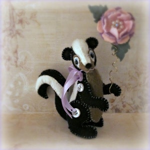 May include: A black and white felt skunk with a purple ribbon around its neck. The skunk is holding a pink felt flower on a wire stem.