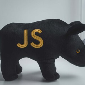 May include: Black plush rhinoceros toy with a white horn and gold embroidered initials "JS".