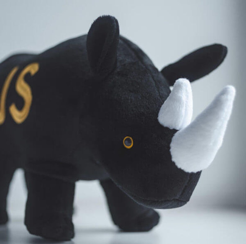 May include: Black plush rhinoceros toy with a white horn and a gold embroidered letter 'S' on its side.