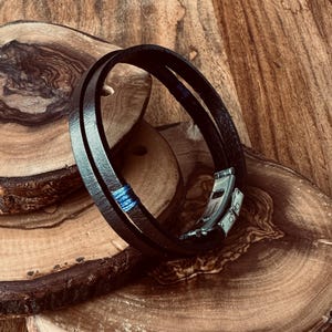 Puede incluir: Un brazalete de cuero negro con detalles de hilo azul y un cierre plateado. El brazalete está envuelto alrededor de una superficie de madera.
