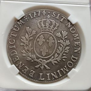 以下が含まれることがあります： 1774年と刻印された銀色の円形コインが、長方形の透明なプラスチック製保護ケースに収められています。コインの中央には王冠を戴いた3つのフルール・ド・リスの紋章が描かれ、周囲には「SIT NOMEN DOMINI BENEDICTUM」という文字が刻まれています。