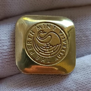 May include: A square gold-coloured metal button with rounded corners. The centre features a circular embossed design of a swan and the text THE PERTH MINT AUSTRALIA. The surface has a polished, reflective finish with a smooth, metallic texture.