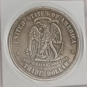 Peut inclure: Une pièce de monnaie en argent United States of America Trade Dollar dans un étui carré en plastique. La pièce représente un aigle tenant des flèches et un bouclier avec le texte IN GOD WE TRUST, ainsi que les inscriptions 420 GRAINS et 900 FINE.