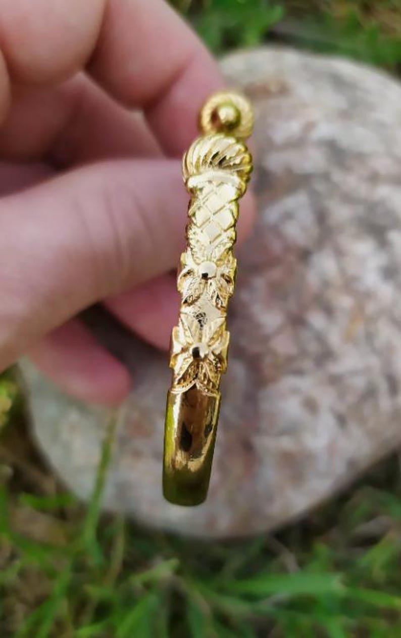 May include: A gold bangle bracelet with a textured design. The bracelet has a floral pattern and a round knob at the top.