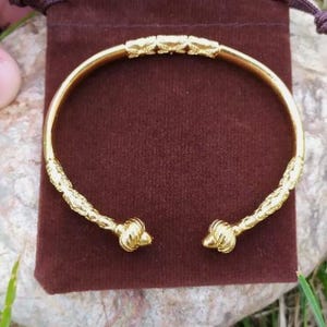 May include: A gold bangle bracelet with a textured design and rounded ends. The bracelet is displayed on a brown velvet pouch.