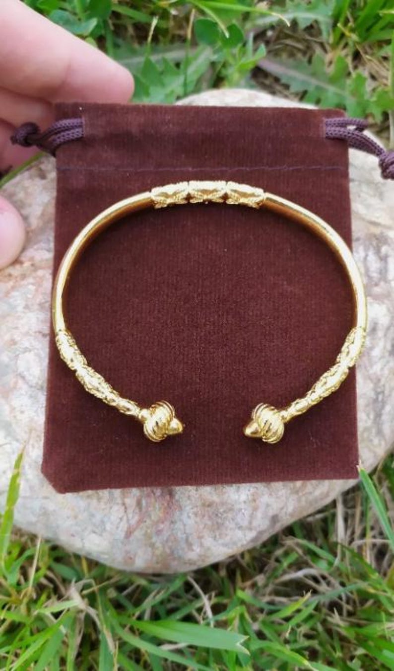 May include: A gold bangle bracelet with a textured design and rounded ends. The bracelet is displayed on a brown velvet pouch.