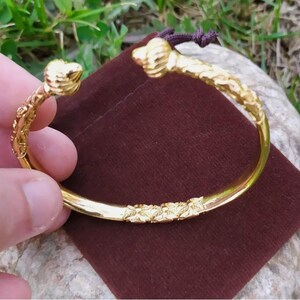 May include: A gold-colored bangle bracelet with a detailed, textured design. The bracelet is open and has a curved shape.