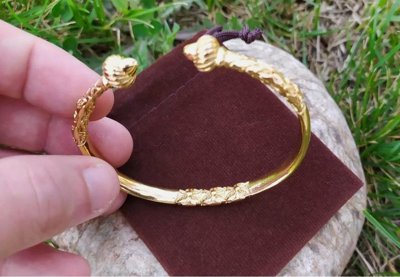 May include: A gold-colored bangle bracelet with a detailed, textured design. The bracelet is open and has a curved shape.