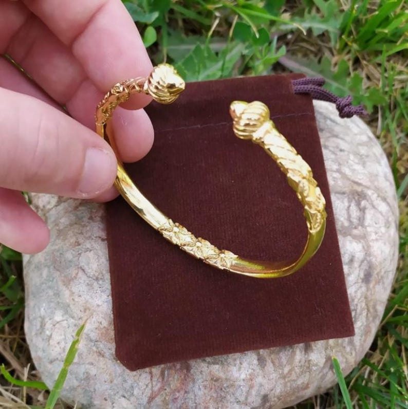 May include: A gold-toned bangle bracelet with a twisted design and floral accents. The ends of the bracelet are capped with decorative, rounded finials.