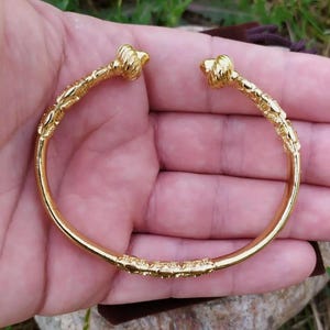 May include: A gold-toned bangle bracelet with a textured design and rounded ends. The bracelet is open and has a simple, elegant style.