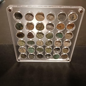 Peut inclure: Un présentoir en acrylique transparent contenant trente-cinq emplacements circulaires, chacun abritant une pièce de monnaie ancienne. Les pièces présentent diverses teintes métalliques, notamment bronze, cuivre et patine verte, disposées en grille.