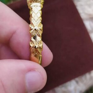 May include: A gold-colored bangle bracelet with a detailed floral design. The bracelet is held in a hand.
