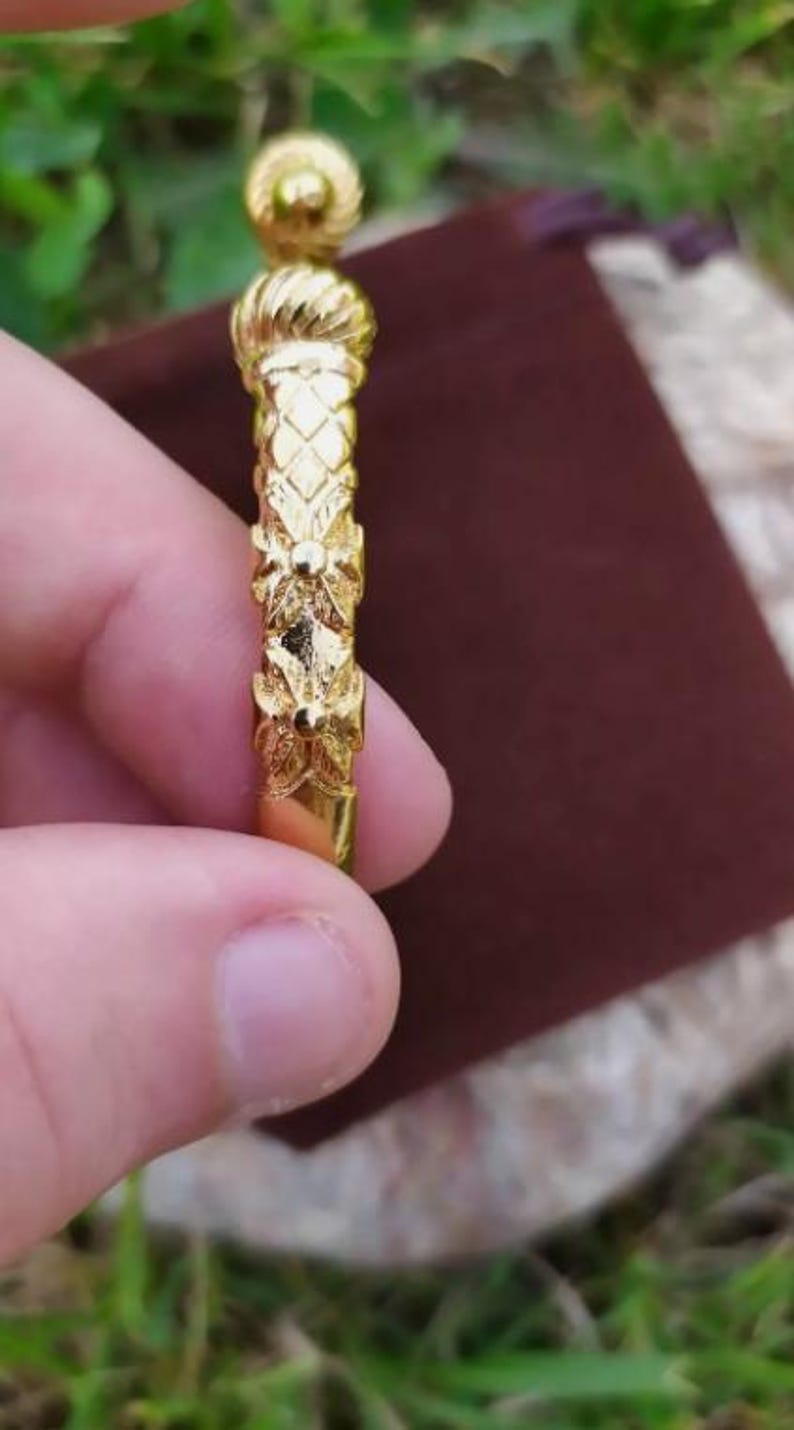 May include: A gold-colored bangle bracelet with a detailed floral design. The bracelet is held in a hand.