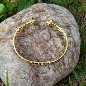 May include: A gold bangle bracelet with a textured design and rounded ends. The bracelet is lying on a rock.