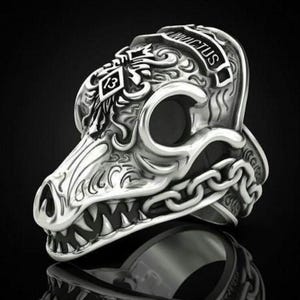 Peut inclure: Une bague en argent avec un motif de crâne détaillé. Le crâne a une chaîne autour de la mâchoire et les mots "INVICTUS" et "13" sont gravés sur le dessus.