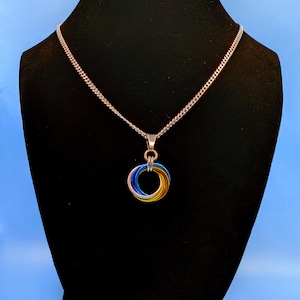 May include: A silver-toned necklace with a chain and a pendant featuring three interlocking rings in blue, yellow, and green. The necklace is displayed on a black mannequin against a blue background.
