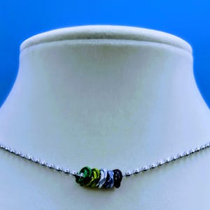 Puede incluir: Un collar de cadena plateada con un dije multicolor hecho de pequeños anillos de metal. El dije es verde, amarillo, blanco y negro.