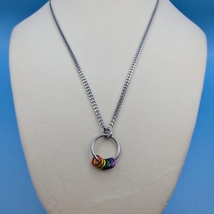 Puede incluir: Un collar de cadena de plata con un colgante de anillo de colores arcoíris. El colgante está hecho de varios anillos pequeños unidos entre sí.