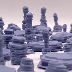 May include: A collection of small, light purple terrain pieces, resembling miniature rock formations and mesas. The pieces vary in height and shape, with layered textures and rounded edges, suitable for tabletop gaming or model building.