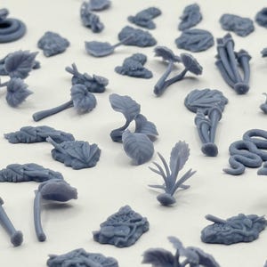 May include: A collection of small, unpainted grey resin miniature figures featuring various botanical elements like leaves, stems, and plants, alongside several coiled snake figures, all arranged in a scattered pattern on a plain surface.