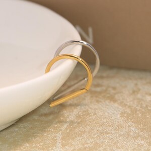 May include: Two U-shaped hoop earrings, one in a polished gold tone and one in a polished silver tone, featuring a smooth, minimalist metal construction with a rounded top and a straight, flat bottom edge, resting against a white curved surface.