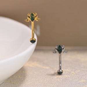 May include: Two curved barbell body jewelry pieces, one gold-toned and one silver-toned. Each features a frog-shaped top ornament with a dark green stone inlay and a matching dark green stone bead at the bottom end of the curved metal post.
