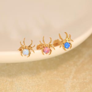 Puede incluir: Tres pendientes de botón con forma de araña en tono dorado, cada uno con una piedra redonda de cabujón. El pendiente izquierdo tiene una piedra blanca, el central una rosa y el derecho una azul. Cada araña tiene ocho patas finas y curvas.
