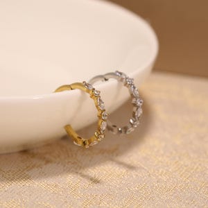 May include: Two hoop earrings, one gold-toned and one silver-toned, each adorned with clear, marquise-cut gemstones. The earrings are displayed on a white surface, with a textured, gold-colored background.