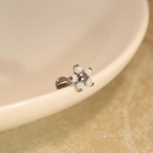 Può includere: Un gioiello da corpo in metallo color argento con una testa a forma di fiore. Il design floreale è composto da cinque piccole pietre rotonde bianco opalescente iridescente disposte attorno a un punto centrale in metallo, fissato su un perno piatto.